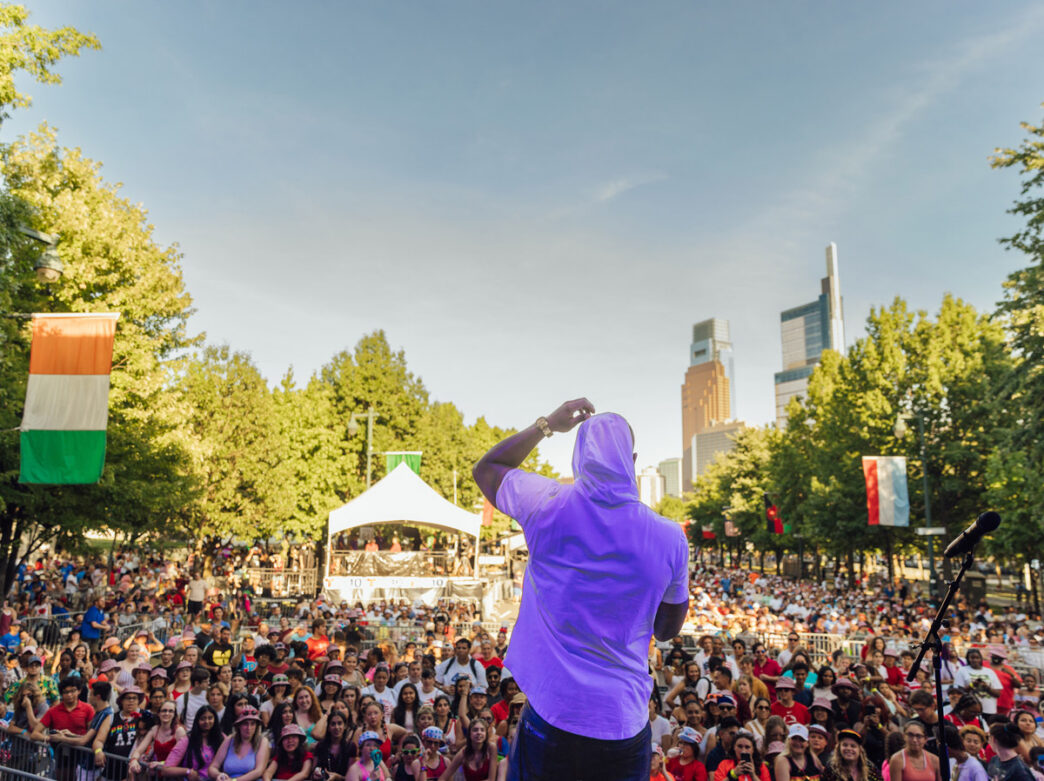 A performer stands on stage facing a large crowd during a July 4th concert on Philadelphia’s Benjamin Franklin Parkway with thousands of attendees filling the tree-lined boulevard and a view of Center City in the background.