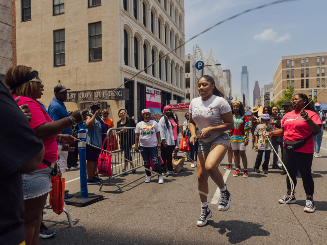 A young woman jumps rope during a double Dutch demonstration at Philadelphia’s Juneteenth Block Party. Spectators line the city street behind barricades as participants turn the ropes and cheer her on, creating an energetic community celebration of Black culture, heritage, and freedom in the heart of downtown Philadelphia.
