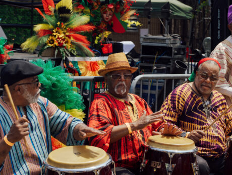 Four men in colorful traditional African-inspired clothing play hand drums on an outdoor stage area during the Juneteenth Festival in Philadelphia. Bright red, green, yellow, and black decorations and floral arrangements fill the background, creating a festive atmosphere as performers celebrate Black culture and heritage through music and community gathering.