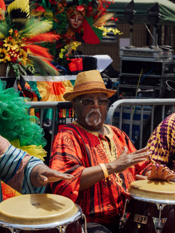 Quatro homens com roupas coloridas e tradicionais de inspiração africana tocam tambores de mão em um palco ao ar livre durante o Juneteenth Festival na Filadélfia. Decorações em vermelho vivo, verde, amarelo e preto e arranjos florais preenchem o fundo, criando uma atmosfera festiva enquanto os artistas celebram a cultura e a herança negra por meio da música e da reunião da comunidade.