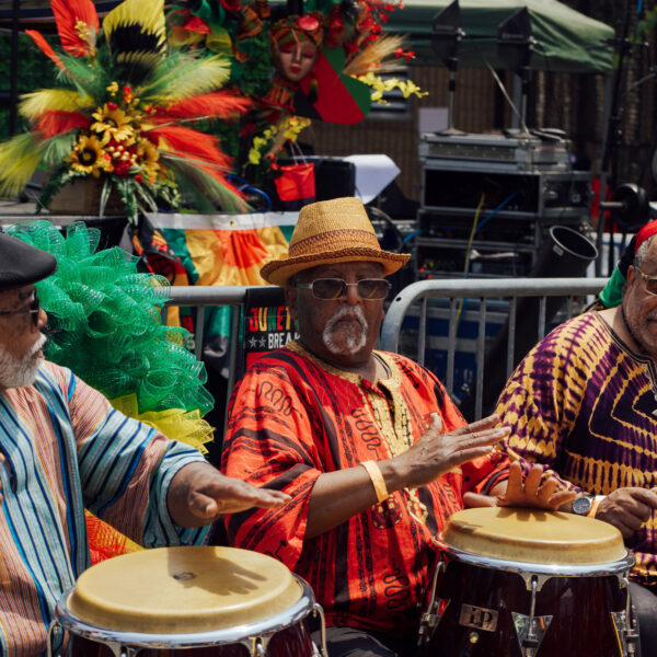 Four men in colorful traditional African-inspired clothing play hand drums on an outdoor stage area during the Juneteenth Festival in Philadelphia. Bright red, green, yellow, and black decorations and floral arrangements fill the background, creating a festive atmosphere as performers celebrate Black culture and heritage through music and community gathering.