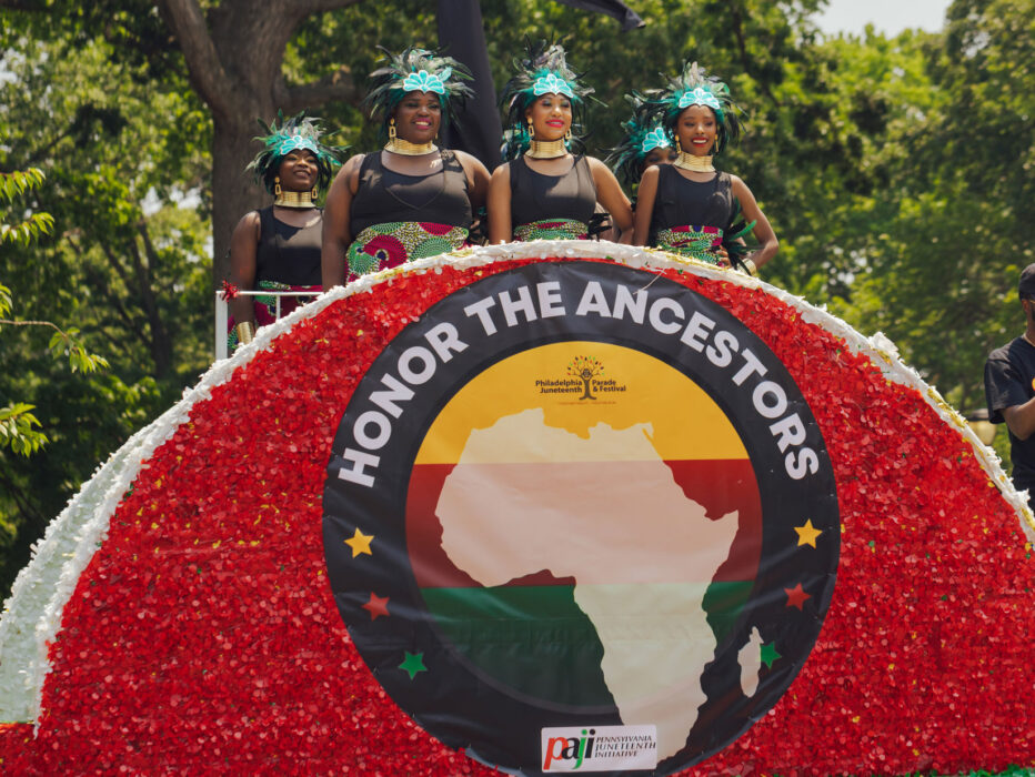 Participants ride on a colorful float during Philadelphia’s Juneteenth Parade. Four women wearing coordinated black outfits, patterned skirts, and feathered headdresses stand atop a red-and-white decorated float displaying a large “Honor the Ancestors” banner featuring the African continent, while parade attendees and trees line the route in the background.