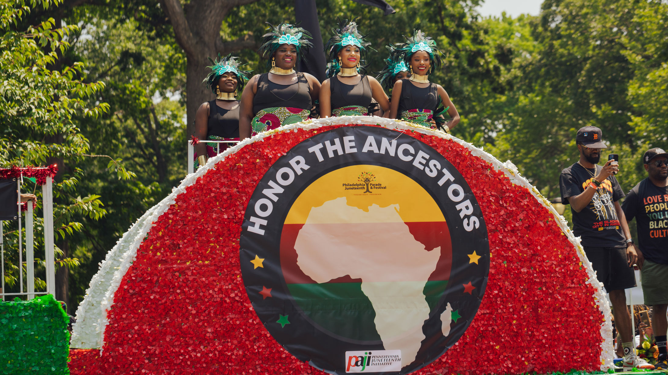 Participants ride on a colorful float during Philadelphia’s Juneteenth Parade. Four women wearing coordinated black outfits, patterned skirts, and feathered headdresses stand atop a red-and-white decorated float displaying a large “Honor the Ancestors” banner featuring the African continent, while parade attendees and trees line the route in the background.