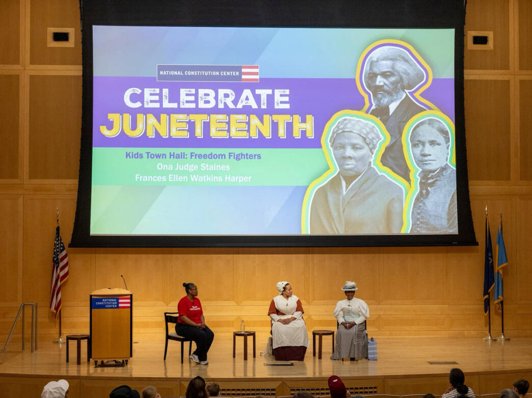 A Juneteenth Town Hall program takes place on the stage of the National Constitution Center in Philadelphia. Three presenters sit before an audience beneath a large screen displaying “Celebrate Juneteenth: Kids Town Hall: Freedom Fighters” alongside historic portraits of abolitionists and civil rights leaders, highlighting educational programming about Black history and freedom movements.