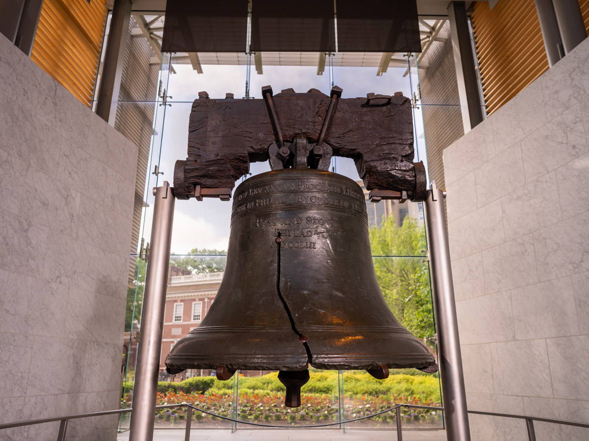 A close-up view of the Liberty Bell inside the Liberty Bell Center in Philadelphia highlights the bell’s famous crack and historic inscription. The bell is displayed in a glass-walled gallery, with Independence Hall and landscaped grounds visible in the background.