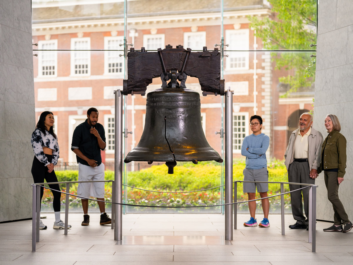 Visitors of different ages stand around the Liberty Bell inside the Liberty Bell Center in Philadelphia, viewing the historic cracked bell from multiple angles. Large windows behind the exhibit frame Independence Hall, connecting two of the nation’s most iconic landmarks.