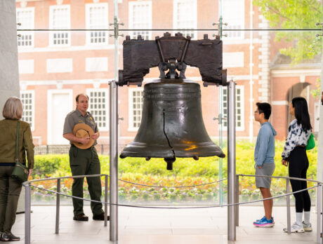 Un guardaparques del Servicio de Parques Nacionales dirige una visita guiada por el interior del Centro Liberty Bell, mientras visitantes de diferentes edades se agrupan alrededor del Liberty Bell para escuchar la explicación. La histórica campana agrietada ocupa un lugar central en la galería, y el Independence Hall se divisa a través de los ventanales que van del piso al techo situados detrás de ella.