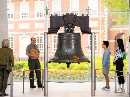 Un guardaparques del Servicio de Parques Nacionales dirige una visita guiada por el interior del Centro Liberty Bell, mientras visitantes de diferentes edades se agrupan alrededor del Liberty Bell para escuchar la explicación. La histórica campana agrietada ocupa un lugar central en la galería, y el Independence Hall se divisa a través de los ventanales que van del piso al techo situados detrás de ella.