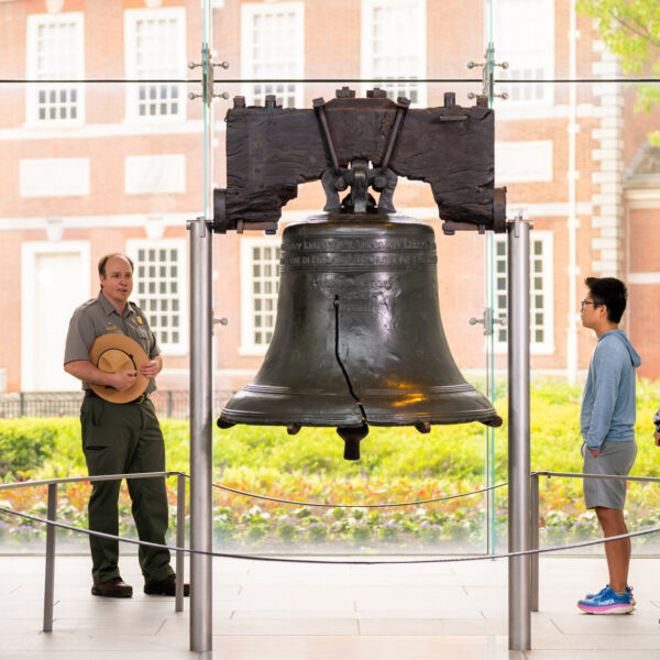 Um guarda florestal do Serviço Nacional de Parques conduz um passeio dentro do Centro Liberty Bell enquanto visitantes de diferentes idades ficam em volta do Liberty Bell ouvindo a apresentação. O histórico sino rachado está centralizado na galeria, com o Independence Hall visível através das janelas do chão ao teto atrás dele.