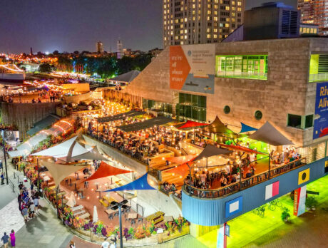 A wide angle shot of an outdoor riverside restaurant, Liberty Point, at night. Overhead lights illuminate the groups and tables