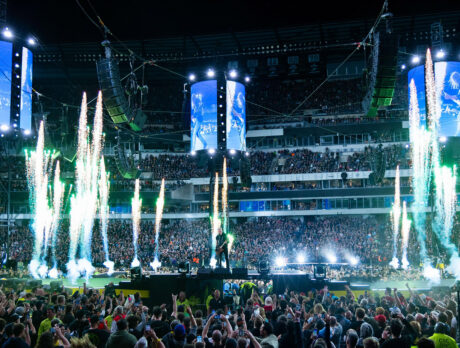 A packed crowd fills Lincoln Financial Field as metal band performs on a circular stadium stage surrounded by towering video screens, dramatic green pyrotechnics and concert lighting.