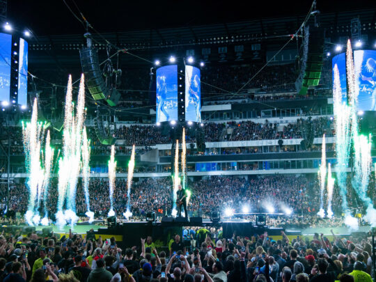 A packed crowd fills Lincoln Financial Field as metal band performs on a circular stadium stage surrounded by towering video screens, dramatic green pyrotechnics and concert lighting.