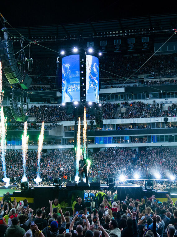 A packed crowd fills Lincoln Financial Field as metal band performs on a circular stadium stage surrounded by towering video screens, dramatic green pyrotechnics and concert lighting.