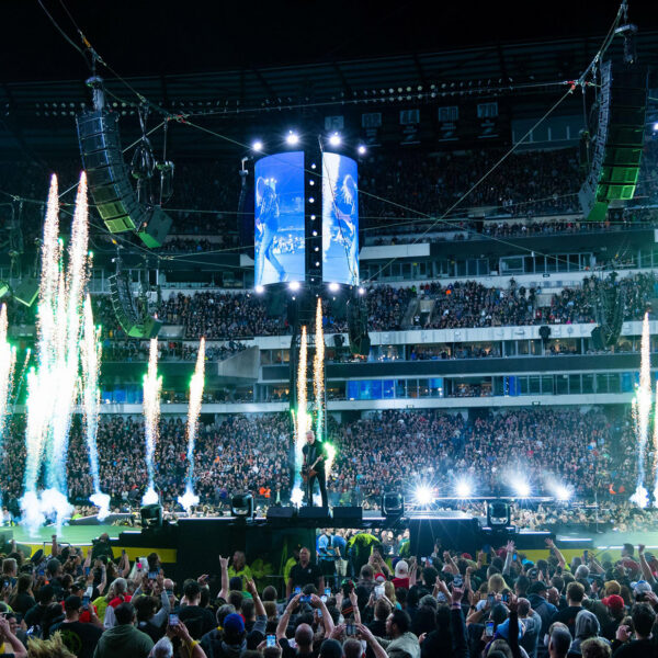 A packed crowd fills Lincoln Financial Field as metal band performs on a circular stadium stage surrounded by towering video screens, dramatic green pyrotechnics and concert lighting.