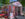 **Texte alternatif :** Une tente d'accueil du Centre d'accueil des visiteurs de Philadelphie, située dans le LOVE Park, présente des visuels colorés « PHILLY 2026 » ainsi que des panneaux indiquant les toilettes, les points d'eau, les plans, les renseignements, la billetterie et la boutique de souvenirs. Des tables et des chaises de café rouges sont disposées à l'extérieur, à l'ombre des arbres, avec les immeubles du centre-ville en arrière-plan.