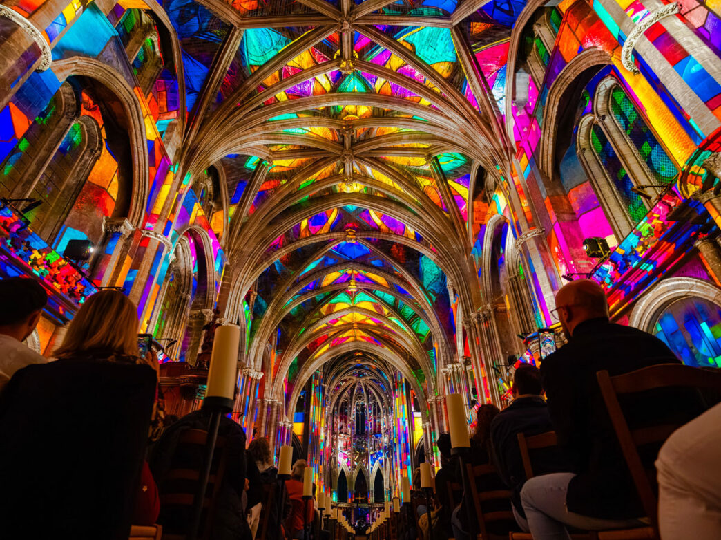 Audience members sit beneath soaring Gothic arches illuminated by vibrant projection-mapped artwork during a Luminescence show. Brilliant colors cover the vaulted ceiling and walls of the historic church, transforming the space into an immersive light-and-sound experience.