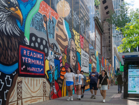 **Alt text:** A group of pedestrians walks along a Center City sidewalk beside a vibrant mural celebrating Philadelphia landmarks, culture and history. The colorful artwork stretches down the block near a bus shelter and modern office buildings.