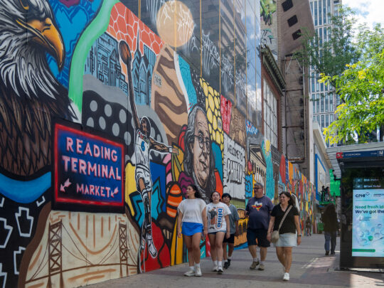 **Alt text:** A group of pedestrians walks along a Center City sidewalk beside a vibrant mural celebrating Philadelphia landmarks, culture and history. The colorful artwork stretches down the block near a bus shelter and modern office buildings.
