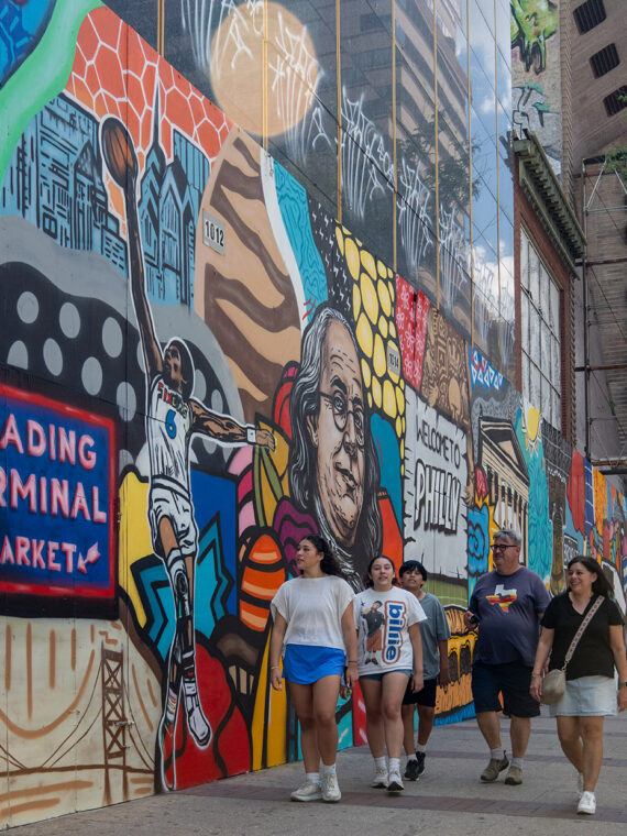 **Alt text:** A group of pedestrians walks along a Center City sidewalk beside a vibrant mural celebrating Philadelphia landmarks, culture and history. The colorful artwork stretches down the block near a bus shelter and modern office buildings.