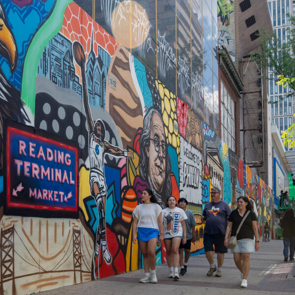**Alt text:** A group of pedestrians walks along a Center City sidewalk beside a vibrant mural celebrating Philadelphia landmarks, culture and history. The colorful artwork stretches down the block near a bus shelter and modern office buildings.