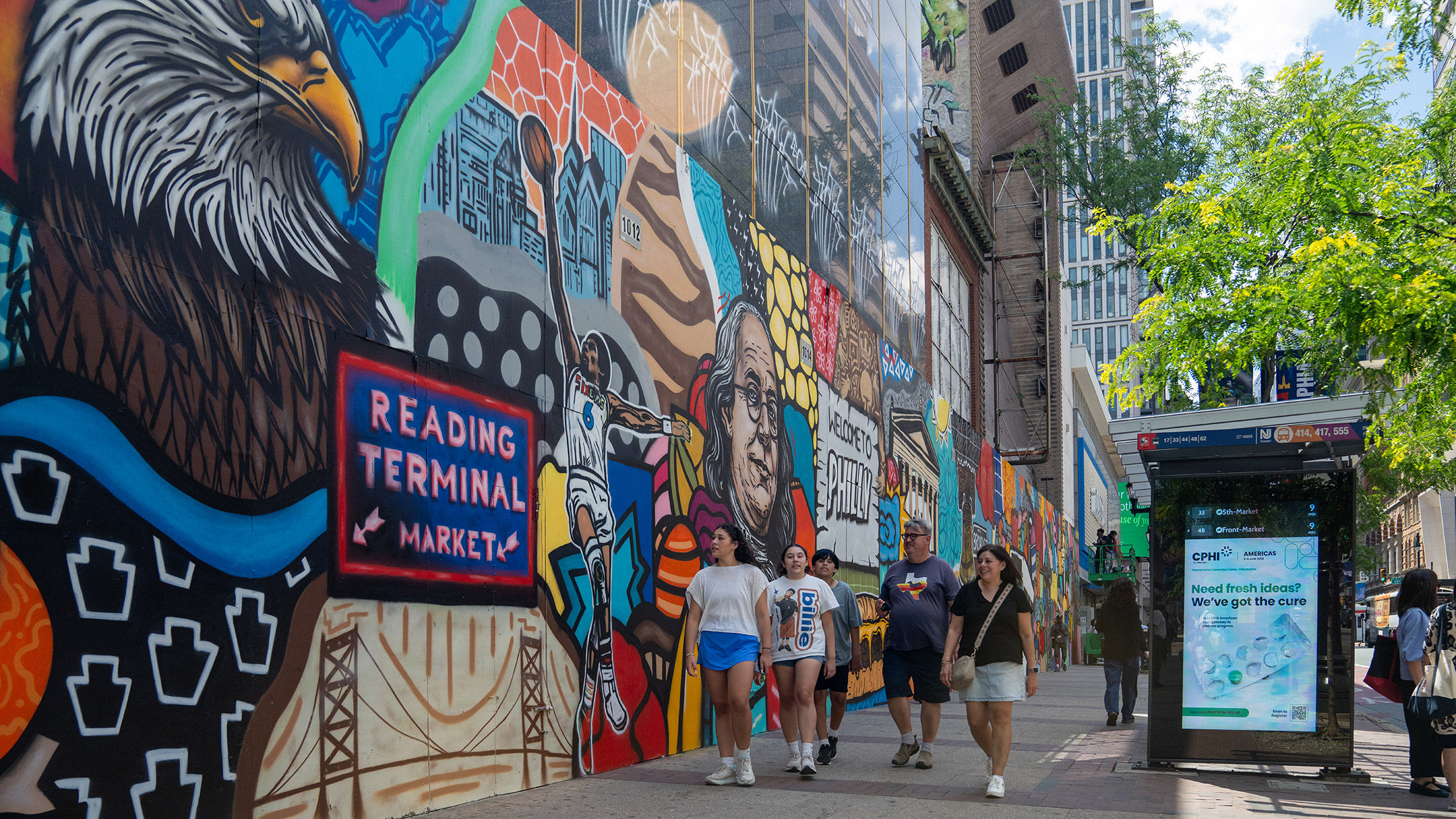 **Alt text:** A group of pedestrians walks along a Center City sidewalk beside a vibrant mural celebrating Philadelphia landmarks, culture and history. The colorful artwork stretches down the block near a bus shelter and modern office buildings.