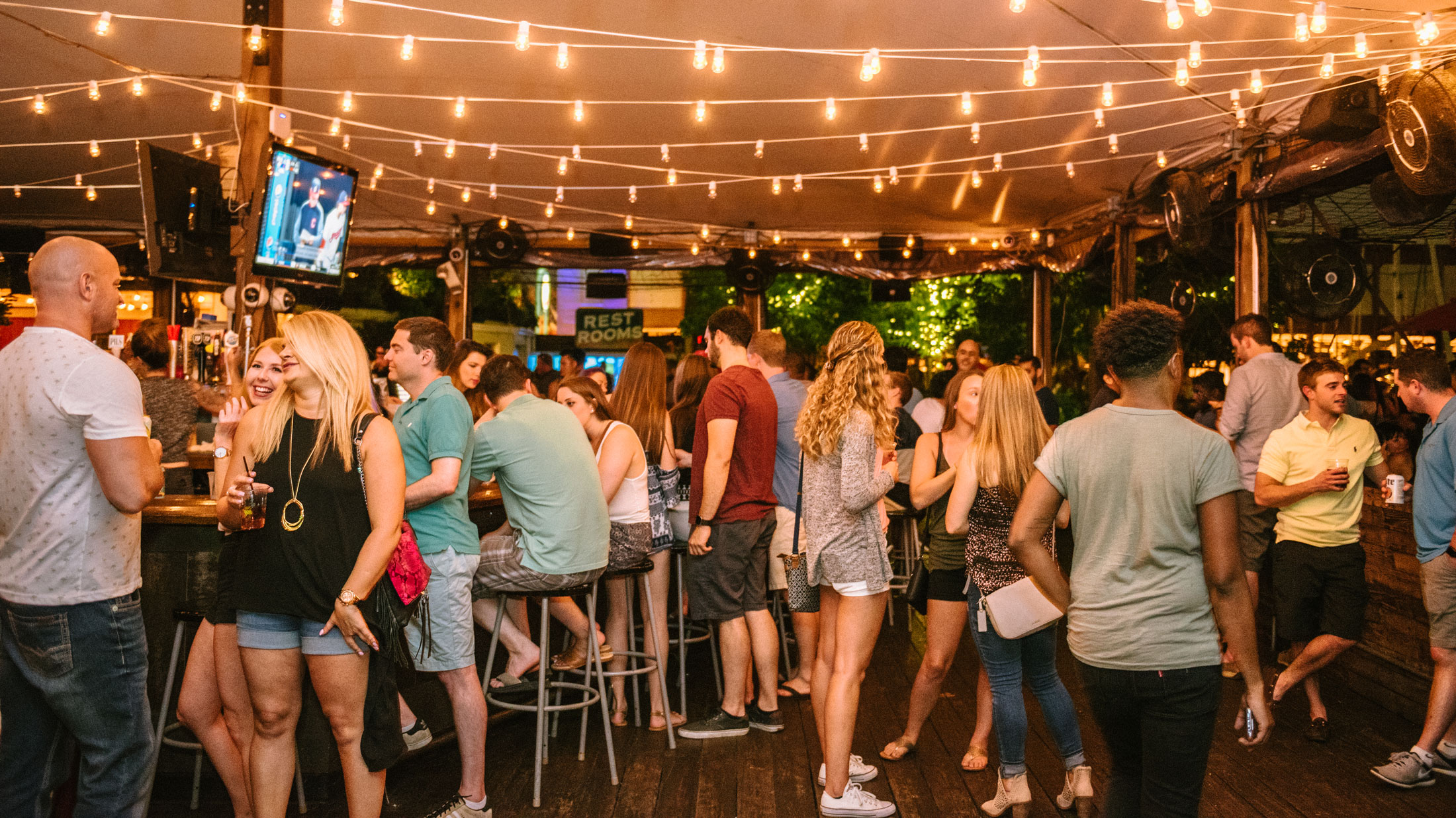 Crowd of patrons socializing and enjoying drinks at Morgan’s Pier in Philadelphia, gathered beneath strings of warm café lights on the waterfront bar’s open-air deck at night.