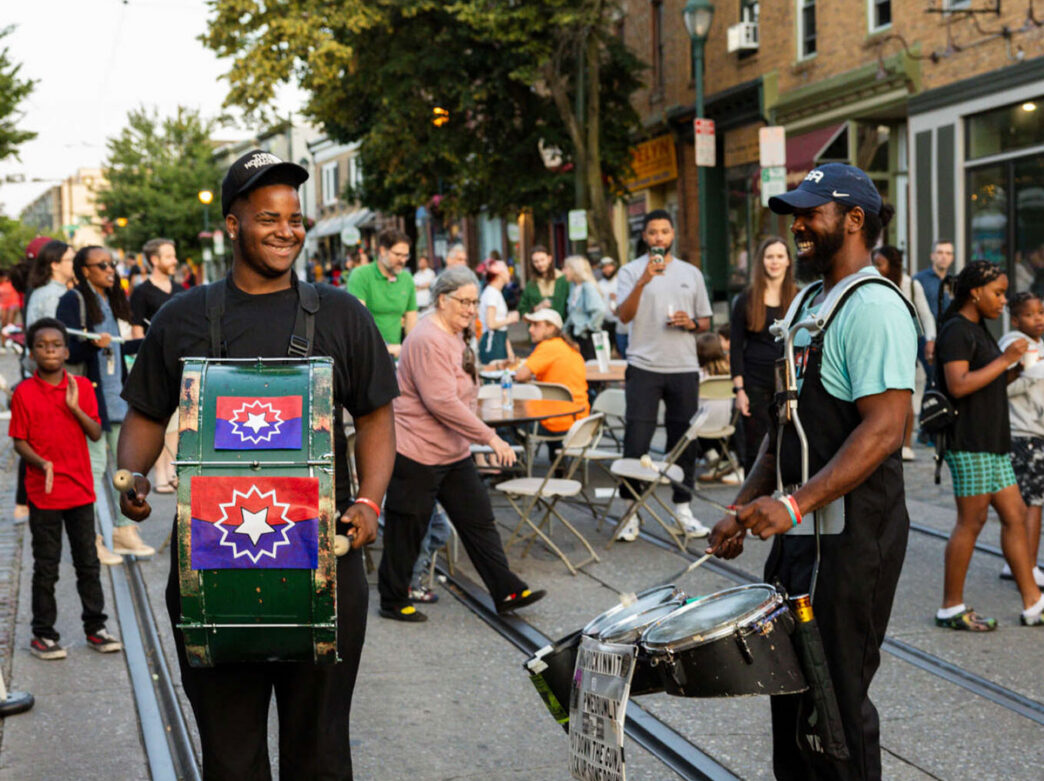 Two drummers smile and perform in the middle of a closed-off street during a Go Mt. Airy Supper Sessions event in Philadelphia. Neighbors gather around outdoor tables and along the sidewalk, creating a lively community atmosphere as evening approaches.