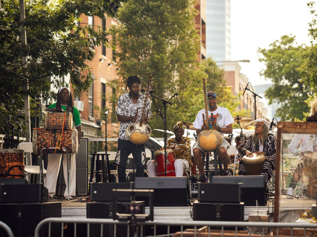 A group of musicians performs at Philadelphia’s ODUNDE Festival on a tree-lined city street. The ensemble plays traditional African instruments, including hand drums and kora harps, while seated and standing across an outdoor stage surrounded by speakers, microphones, and festival crowds enjoying the live music.