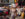 A performer in white ceremonial attire dances in the middle of a crowded street during Philadelphia’s ODUNDE Festival. Wearing a white headwrap, shell jewelry, and body paint, she extends a bundle of wooden staffs while spectators gather around to watch, photograph, and cheer. Drummers playing conga-style drums accompany the performance, creating a vibrant atmosphere that celebrates African and African American culture and traditions.