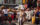 A performer in white ceremonial attire dances in the middle of a crowded street during Philadelphia’s ODUNDE Festival. Wearing a white headwrap, shell jewelry, and body paint, she extends a bundle of wooden staffs while spectators gather around to watch, photograph, and cheer. Drummers playing conga-style drums accompany the performance, creating a vibrant atmosphere that celebrates African and African American culture and traditions.