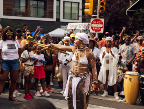 A performer in white ceremonial attire dances in the middle of a crowded street during Philadelphia’s ODUNDE Festival. Wearing a white headwrap, shell jewelry, and body paint, she extends a bundle of wooden staffs while spectators gather around to watch, photograph, and cheer. Drummers playing conga-style drums accompany the performance, creating a vibrant atmosphere that celebrates African and African American culture and traditions.