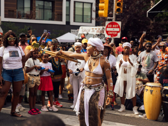 A performer in white ceremonial attire dances in the middle of a crowded street during Philadelphia’s ODUNDE Festival. Wearing a white headwrap, shell jewelry, and body paint, she extends a bundle of wooden staffs while spectators gather around to watch, photograph, and cheer. Drummers playing conga-style drums accompany the performance, creating a vibrant atmosphere that celebrates African and African American culture and traditions.