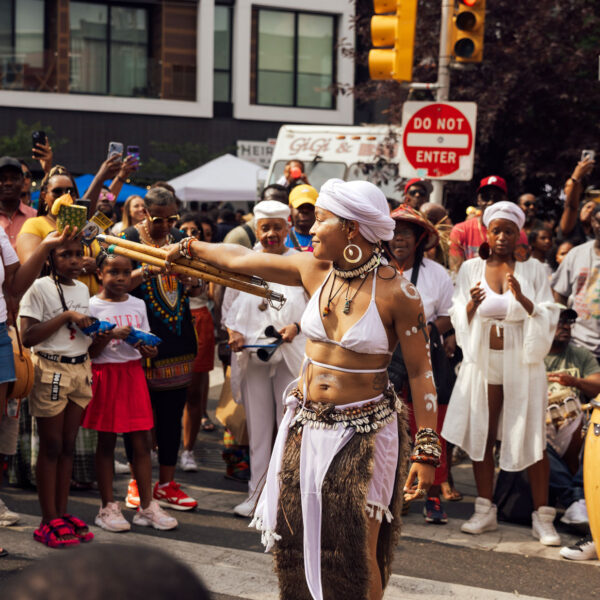 Uma artista em trajes cerimoniais brancos dança no meio de uma rua lotada durante o ODUNDE Festival da Filadélfia. Usando um envoltório branco na cabeça, joias de concha e pintura corporal, ela estende um feixe de cajados de madeira enquanto os espectadores se reúnem para assistir, fotografar e aplaudir. Bateristas tocando tambores estilo conga acompanham a performance, criando uma atmosfera vibrante que celebra a cultura e as tradições africanas e afro-americanas.