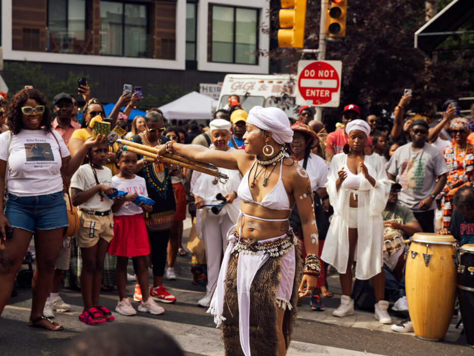 A performer in white ceremonial attire dances in the middle of a crowded street during Philadelphia’s ODUNDE Festival. Wearing a white headwrap, shell jewelry, and body paint, she extends a bundle of wooden staffs while spectators gather around to watch, photograph, and cheer. Drummers playing conga-style drums accompany the performance, creating a vibrant atmosphere that celebrates African and African American culture and traditions.