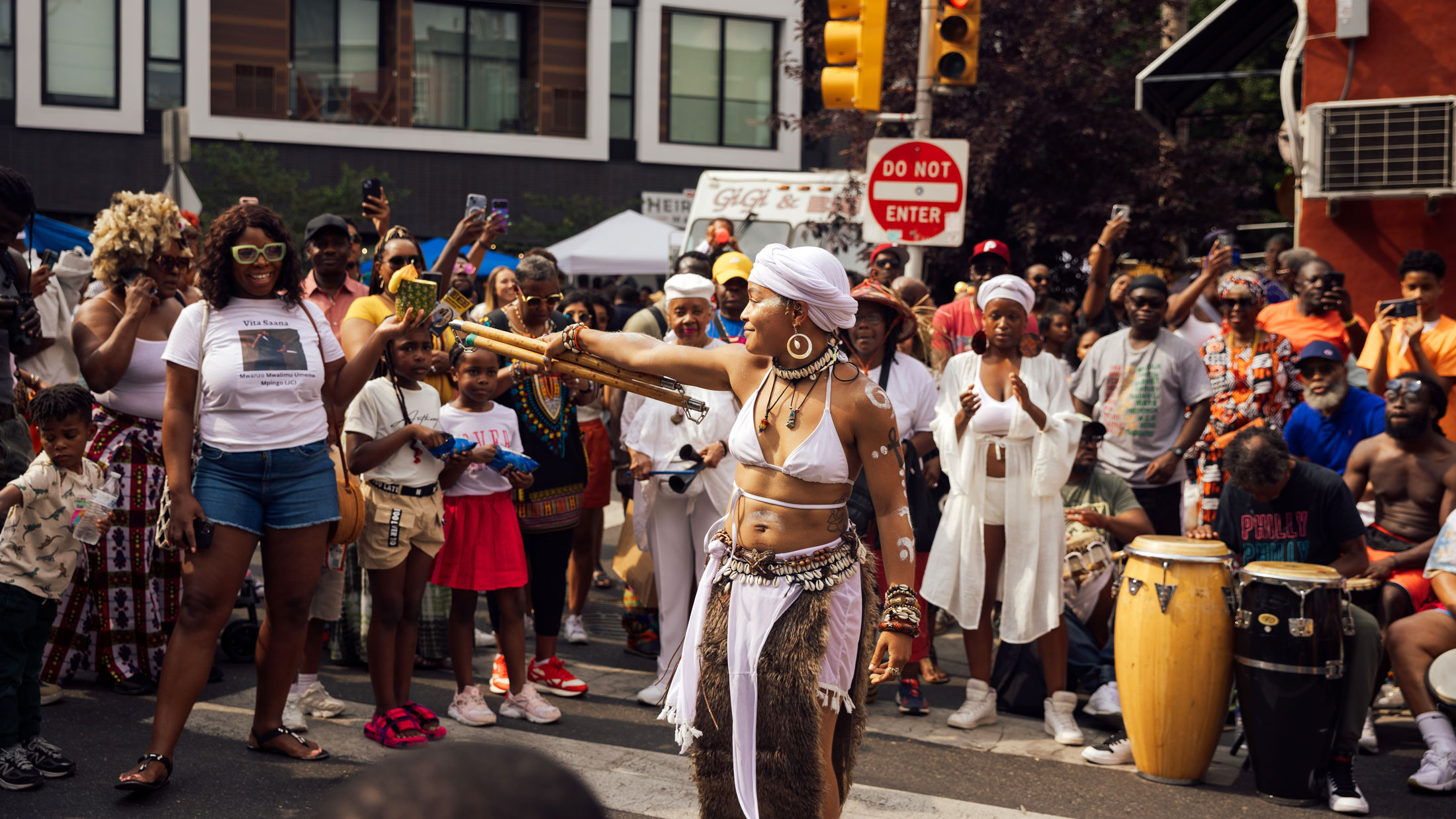 A performer in white ceremonial attire dances in the middle of a crowded street during Philadelphia’s ODUNDE Festival. Wearing a white headwrap, shell jewelry, and body paint, she extends a bundle of wooden staffs while spectators gather around to watch, photograph, and cheer. Drummers playing conga-style drums accompany the performance, creating a vibrant atmosphere that celebrates African and African American culture and traditions.
