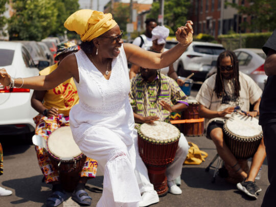 Una mujer con un vestido blanco y un pañuelo amarillo en la cabeza baila alegremente en medio de una calle durante el Festival ODUNDE de Filadelfia, rodeada de percusionistas que tocan tambores de mano. Detrás de ella actúan participantes del festival vestidos con ropa de coloridos estampados, mientras que los autos estacionados y las casas adosadas bordean la calle del barrio, bañada por el sol.