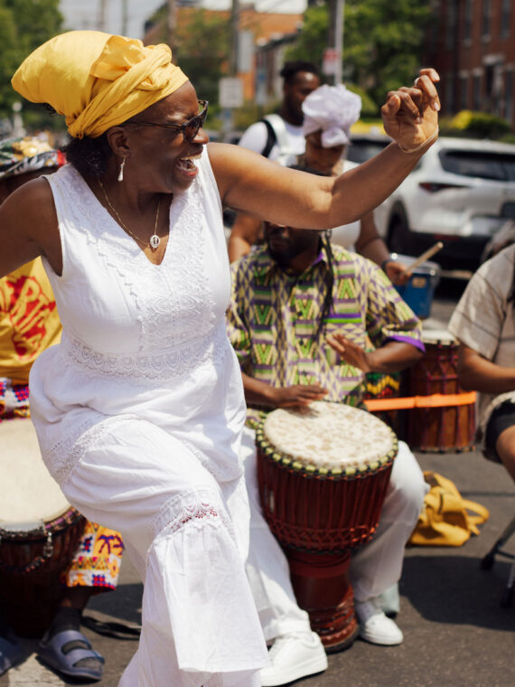 A woman in a white dress and yellow headwrap dances joyfully in the middle of a street during Philadelphia’s ODUNDE Festival, surrounded by drummers playing hand drums. Festival participants in colorful patterned clothing perform behind her, while parked cars and rowhouses line the sunlit neighborhood street.