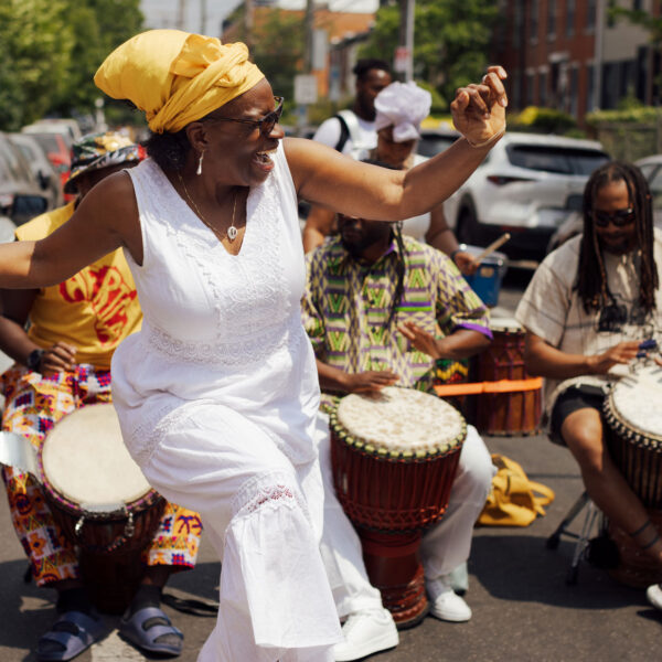 Una mujer con un vestido blanco y un pañuelo amarillo en la cabeza baila alegremente en medio de una calle durante el Festival ODUNDE de Filadelfia, rodeada de percusionistas que tocan tambores de mano. Detrás de ella actúan participantes del festival vestidos con ropa de coloridos estampados, mientras que los autos estacionados y las casas adosadas bordean la calle del barrio, bañada por el sol.