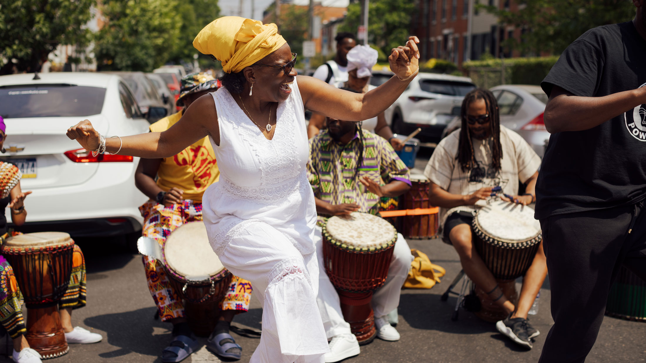 Une femme vêtue d'une robe blanche et d'un bandeau jaune danse joyeusement au milieu d'une rue pendant le festival ODUNDE de Philadelphie, entourée de batteurs jouant des tambours à main. Des participants au festival, vêtus de vêtements à motifs colorés, se produisent derrière elle, tandis que des voitures garées et des maisons en rangée bordent la rue de quartier éclairée par le soleil.