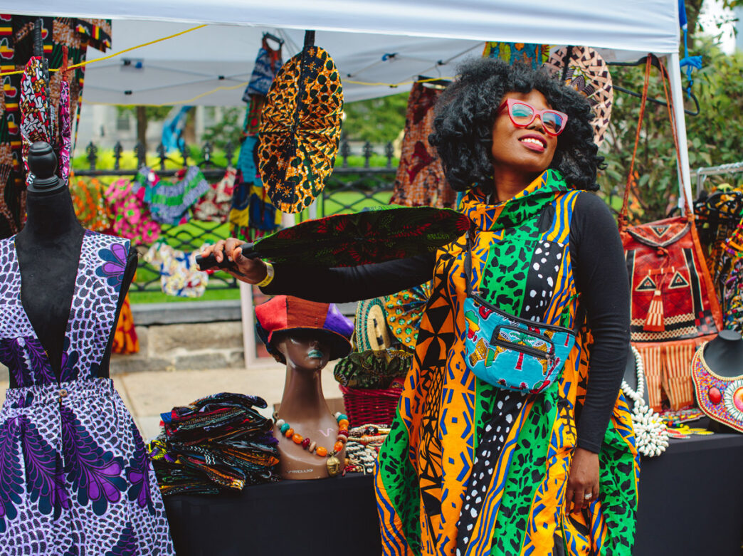 At Philadelphia’s ODUNDE Festival, a smiling woman wearing a vibrant green, yellow, orange, and black patterned dress poses with a decorative fan at an outdoor market stall. The booth is filled with colorful African-inspired clothing, accessories, handbags, jewelry, and textiles displayed under a white canopy.
