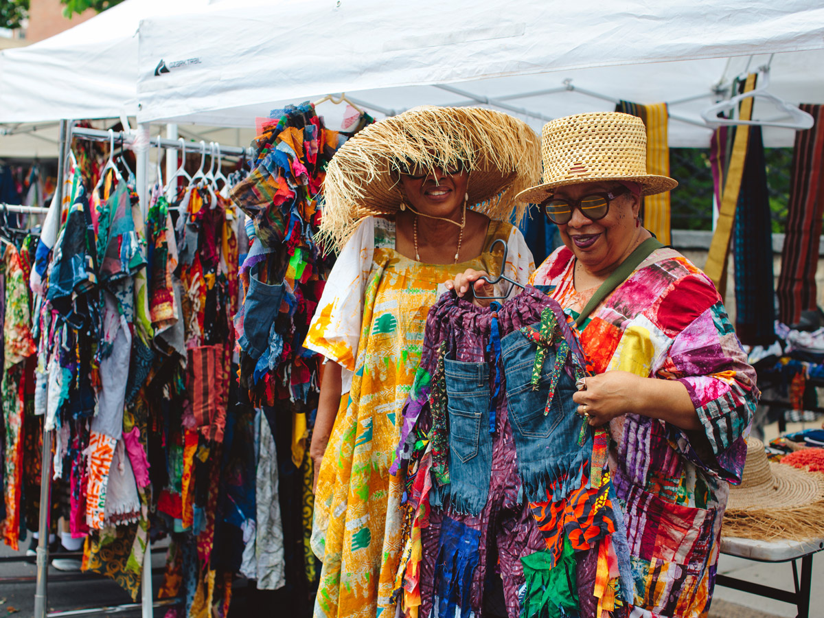 At Philadelphia’s ODUNDE Festival, two women in patterned dresses and woven sun hats examine a vibrant patchwork garment at an outdoor market stall. Clothing racks filled with colorful handmade apparel and textiles surround them beneath white canopy tents, highlighting the festival’s lively marketplace atmosphere.