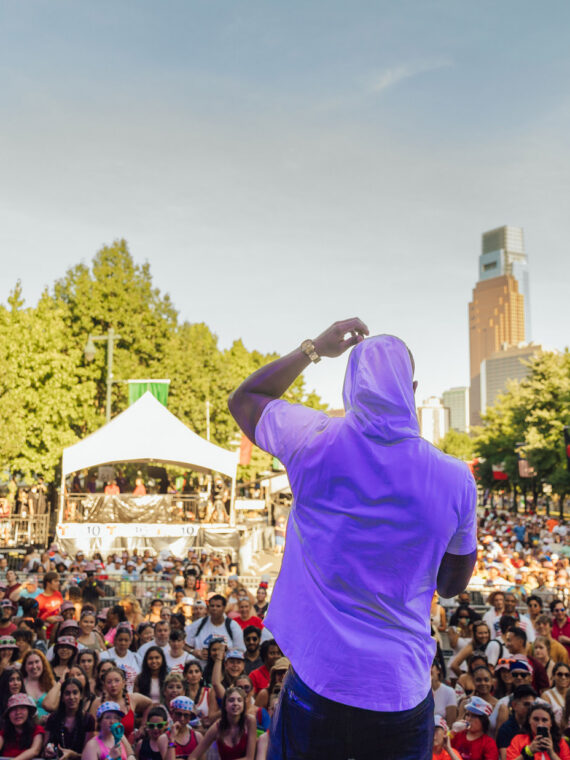 A performer stands on stage facing a large crowd during a July 4th concert on Philadelphia’s Benjamin Franklin Parkway with thousands of attendees filling the tree-lined boulevard and a view of Center City in the background.