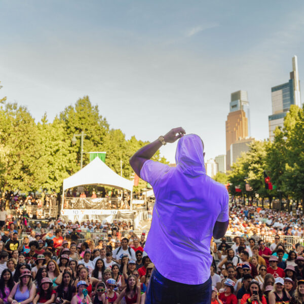 Un artista se encuentra en el escenario frente a una gran multitud durante un concierto del 4 de julio en el Benjamin Franklin Parkway de Filadelfia, con miles de asistentes que llenan el bulevar arbolado y el centro de la ciudad como telón de fondo.