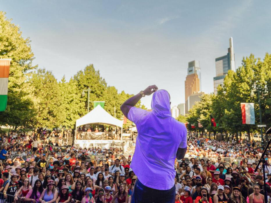 Tijdens een concert op 4 juli op de Benjamin Franklin Parkway in Philadelphia staat een artiest op het podium met het gezicht naar een groot publiek, terwijl duizenden bezoekers de met bomen omzoomde boulevard vullen en op de achtergrond het stadscentrum te zien is.