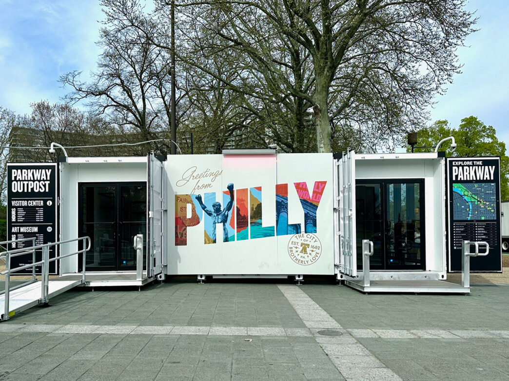 **Texte alternatif :** Le centre d'accueil “ Parkway Outpost ”, situé sur le Benjamin Franklin Parkway à Philadelphie, présente une grande fresque murale intitulée « Greetings from Philly », flanquée de panneaux d'information destinés aux visiteurs et d'entrées accessibles, avec des arbres qui s'élèvent derrière ce bâtiment moderne.