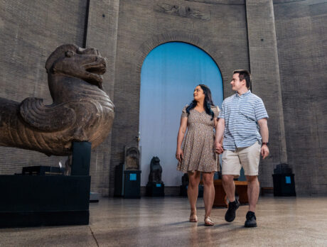 Inside the Penn Museum in Philadelphia, a couple strolls through a spacious gallery while admiring ancient artifacts. A massive stone sculpture resembling a mythological guardian figure dominates the foreground, with additional historic statues and architectural details visible throughout the museum’s grand interior.