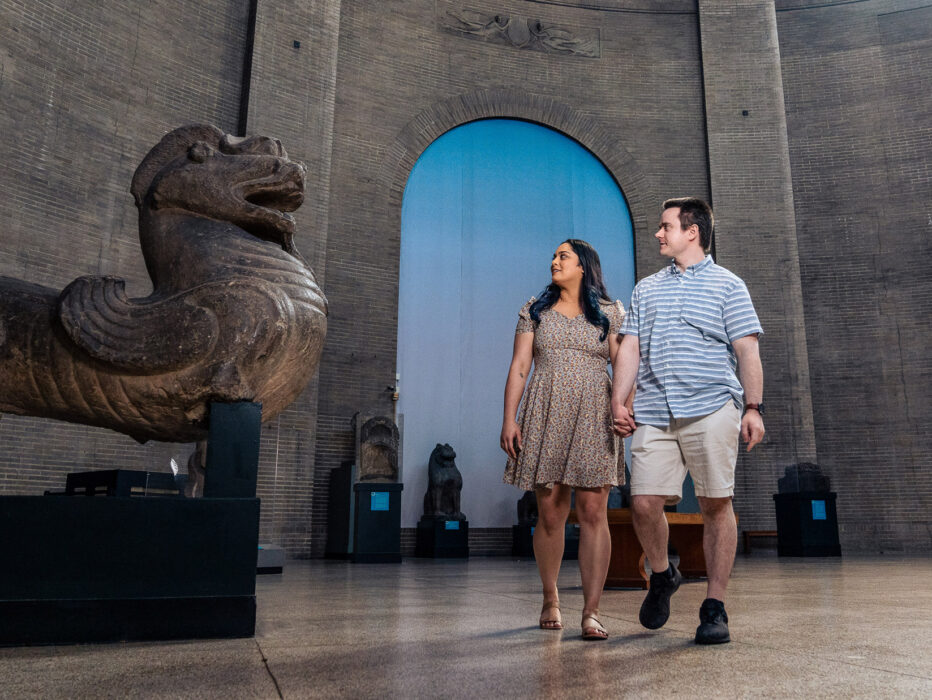 Inside the Penn Museum in Philadelphia, a couple strolls through a spacious gallery while admiring ancient artifacts. A massive stone sculpture resembling a mythological guardian figure dominates the foreground, with additional historic statues and architectural details visible throughout the museum’s grand interior.