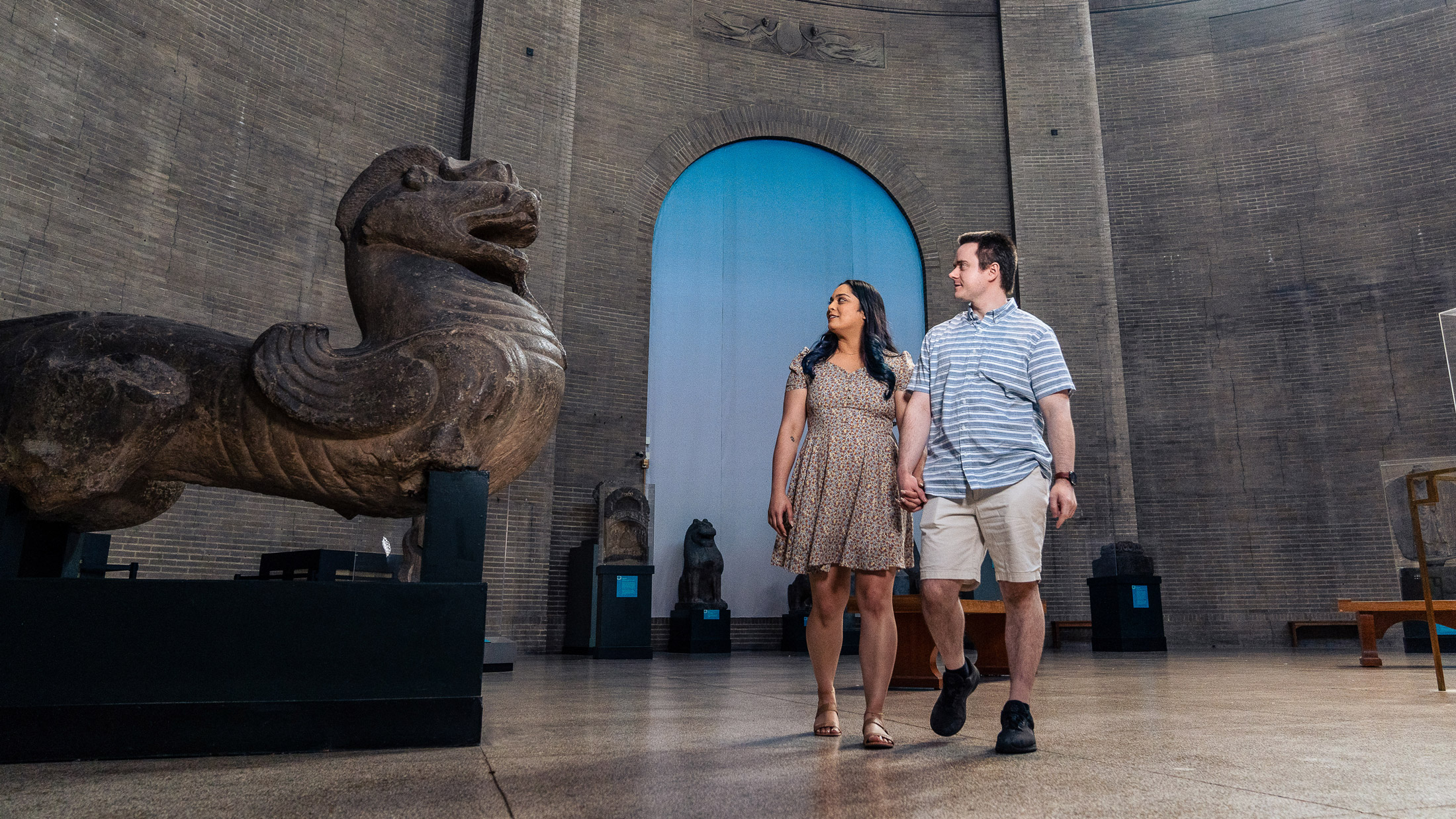 À l'intérieur du Penn Museum de Philadelphie, un couple se promène dans une galerie spacieuse tout en admirant des objets anciens. Une sculpture massive en pierre ressemblant à un gardien mythologique domine le premier plan, tandis que d'autres statues historiques et détails architecturaux sont visibles dans l'intérieur grandiose du musée.