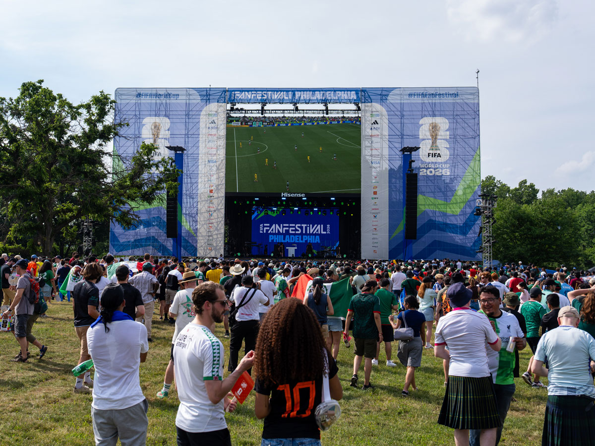 Una gran multitud se reúne en un campo de césped en el Festival de Aficionados de la FIFA de Filadelfia para ver un partido de fútbol en una pantalla gigante instalada sobre un escenario tipo concierto. Los aficionados, vestidos con camisetas y portando banderas nacionales, llenan el recinto al aire libre, decorado con motivos de la Copa Mundial de la FIFA 2026 y rodeado de árboles.