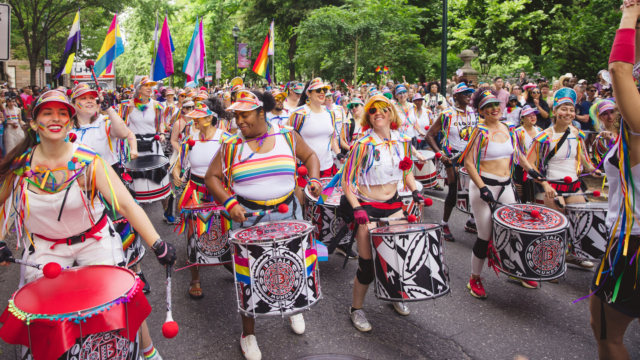 A large, diverse drumline marches in the Philly Pride March, playing colorful drums while wearing rainbow-themed outfits, face paint, and visors. Pride flags, including rainbow, transgender, and nonbinary flags, line the tree-shaded street as crowds of spectators cheer from both sides, creating a festive and energetic atmosphere.